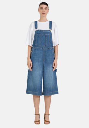 Donna che indossa una camicia bianca a maniche corte sotto un paio di pantaloncini denim a gamba larga con bretelle e sandali marroni con tacco, in piedi davanti a uno sfondo semplice.