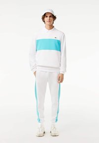 Chándal blanco con una franja horizontal turquesa en el pulóver y paneles laterales a juego en los pantalones. Cuenta con un cuello y puños.