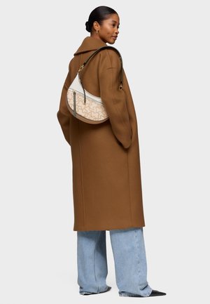 Donna che indossa un lungo cappotto marrone, jeans azzurri, scarpe nere a punta e una borsa beige con fantasia e tracolla nera.