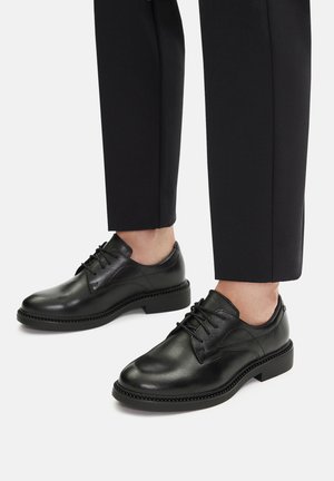 Zapatos de vestir de cuero negro con un acabado suave, que cuentan con cierre de cordones y detalles de costura sutiles, combinados con pantalones negros ajustados.