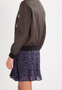 Giacca bomber marrone scuro con vita aderente e tasca con zip sul maniche, indossata sopra un vestito skater floreale blu navy con orlo fluido.