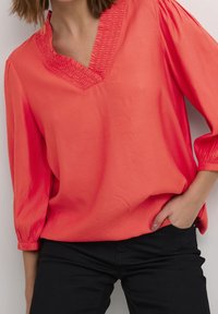 Blusa corallo con scollatura a V e dettagli arricciati sul collo. Maniche a tre quarti con polsini arricciati. Texture morbida e liscia.