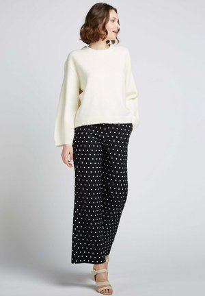 Un pull crème surdimensionné avec des poignets côtelés associé à un pantalon noir à pois larges. Des sandales beige à talons légers complètent l'ensemble.