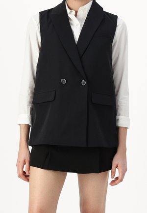 Personne portant un gilet noir sans manches à double boutonnage sur une chemise blanche à manches longues avec les poignets retroussés et une jupe noire.