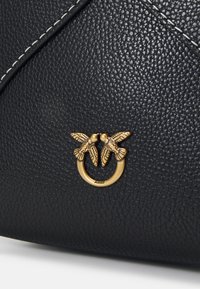 Czarna torebka skórzana o teksturowanej powierzchni, z złotym zapięciem w kształcie dwóch ptaków, z logo marki pod spodem oraz kontrastowym białym przeszyciem.