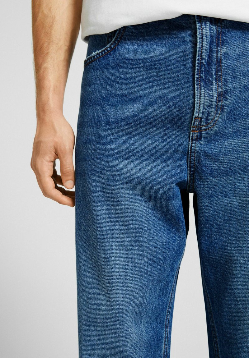 Jeans de mezclilla azul con corte recto, que presentan costuras naranjas, diseño de cinco bolsillos y una textura suave. Mano descansando sobre la cadera.