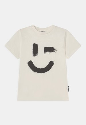T-shirt crème à manches courtes avec un motif visage souriant clignant de l'œil réalisé au pinceau noir sur le devant et une petite étiquette en tissu sur le côté.