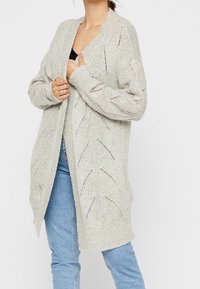 Person wearing a light gray open-knit long cardigan over a black top and blue jeans, holding the cardigan near the chest.