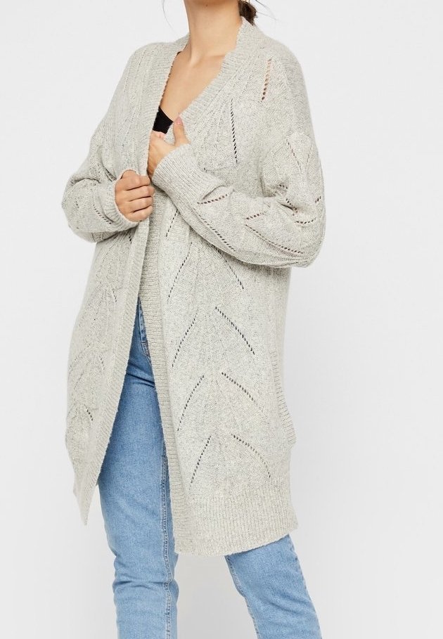 Person wearing a light gray open-knit long cardigan over a black top and blue jeans, holding the cardigan near the chest.