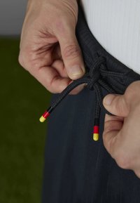 Black drawstring waistband with hand gripping the cord, featuring colored plastic tips in red, yellow, and black. Smooth fabric texture.