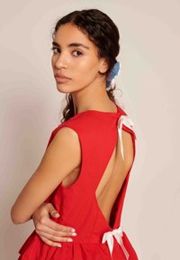 Jovem mulher vestindo um vestido vermelho sem mangas, com as costas abertas e atado com laços brancos, olhando por cima do ombro contra um fundo simples.