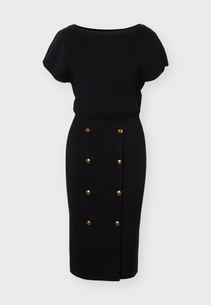 Femme portant un haut noir à manches courtes, une jupe noire boutonnée aux genoux avec des boutons dorés et des bottines noires, debout devant un fond clair.