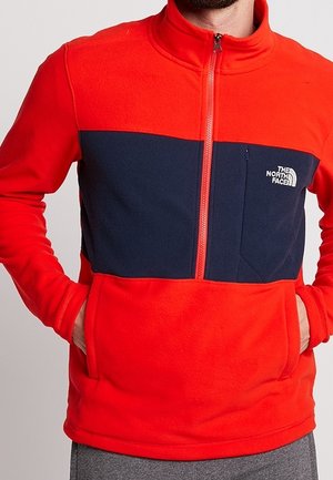 Czerwona i granatowa kurtka polarowa z zamkiem błyskawicznym i kieszenią na piersi, ręce w przednich kieszeniach, z logo The North Face na piersi.