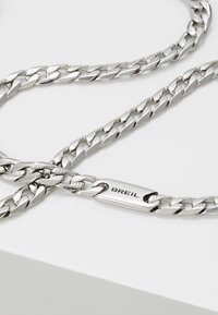 Hopeaketju, joka on valmistettu hopeanvärisestä metallista, jossa on toisiinsa kytkettyjä, litteitä lenkkejä ja suorakulmainen lukko, johon on kaiverrettu "BREIL". Pintakäytävä on sileä ja heijastava.