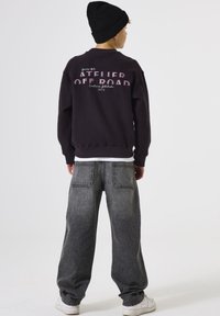 Sudadera negra con gráfico de "ATELIER OFFROAD", combinada con vaqueros grises deslavados de corte holgado y zapatillas blancas. La persona lleva un gorro negro.