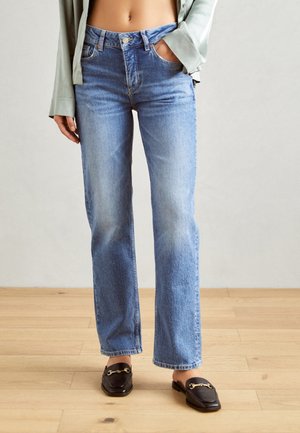 Lys blå høyt liv jeans med rette ben og falmet tekstur, kombinert med svarte loafers med en gullkjede detaljer.