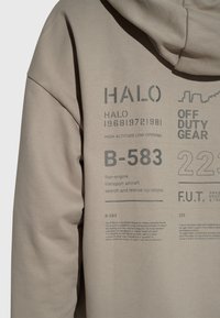 Zbliżenie beżowej bluzy z nadrukiem tekstu, w tym "HALO," "OFF DUTY GEAR," "B-583" oraz opis czterosilnikowego samolotu transportowego.