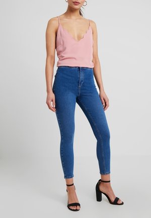 Mujer con top tipo camisola rosa con borde ondulado, jeans ajustados azul de talle alto y tacones negros con tira en el tobillo, de pie contra un fondo liso.