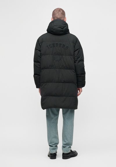 Homme portant une longue doudoune noire avec une capuche, un jean bleu clair et des chaussures noires, debout de dos sur un fond uni.