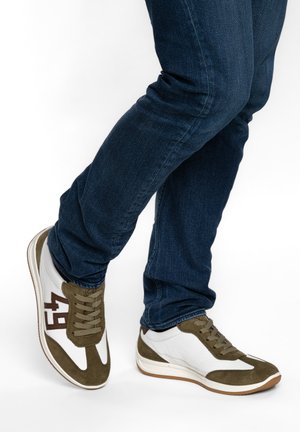 Person, der blaue Jeans und weiße Turnschuhe mit olivgrünen Akzenten und der Zahl 49 trägt, steht mit überkreuzten Beinen.