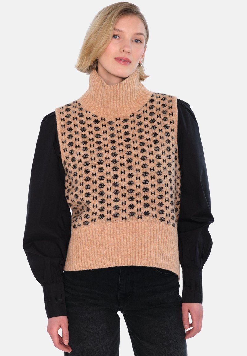 Strikket ermeløs genser med høy hals i ferskenfarge med svarte geometriske mønstre, lagd over en svart langermet skjorte, med ribbede detaljer.