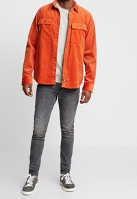 Camisa de pana naranja con dos bolsillos en el pecho, cierre de botones y un suéter gris debajo, combinada con jeans grises y zapatillas oscuras.