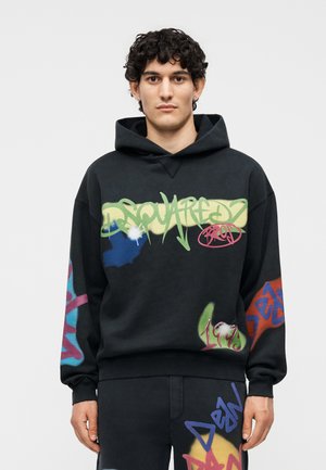 Νέος άνδρας που φοράει μαύρη φούτερ με κουκούλα και παντελόνι με πολύχρωμο κείμενο σε στιλ γκράφιτι και αφηρημένα σχέδια μπροστά και στα μανίκια.