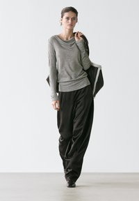 Femme marchant à l'intérieur, portant un haut à manches longues gris, un pantalon noir ample, des chaussures noires et portant une veste noire sur l'épaule.