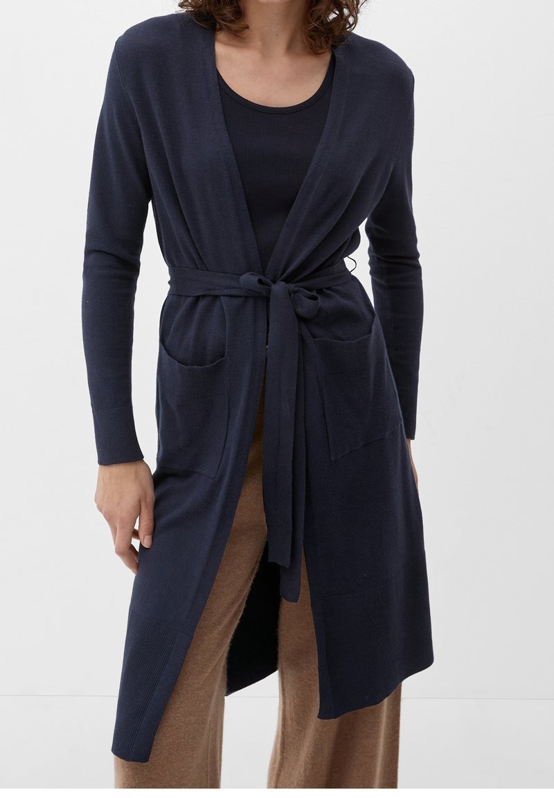 Femme portant un cardigan bleu marine ceinturé avec des poches devant, sur un haut noir et un pantalon marron, sur un fond clair uni.