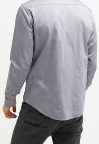 Camiseta gris de manga larga con textura suave, que presenta un puño con botones y un dobladillo curvado. Vista trasera muestra un diseño simple y minimalista.