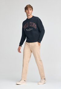 Marinblå sweatshirt med "POLO CLUB" i fet kastanjefärgad text, kombinerad med beige byxor och ljusa sneakers, modern passform, avslappnad stil.