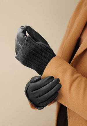 Person, die schwarze Leder- und Strick-Winterhandschuhe trägt und den Ärmel eines mantel in Kamelhaarfarbe anpasst.