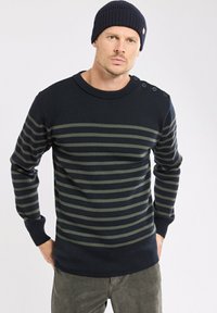 Homme portant un bonnet bleu marine et un pull bleu marine avec des rayures horizontales vertes et des détails de bouton sur l'épaule, debout les mains dans les poches.