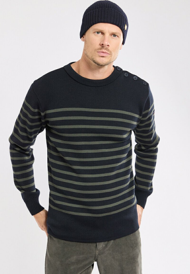 Homme portant un bonnet bleu marine et un pull bleu marine avec des rayures horizontales vertes et des détails de bouton sur l'épaule, debout les mains dans les poches.