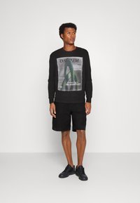 Czarna bluza z grafiką inspirowaną sztuką na przodzie, zestawiona z czarnymi szortami i czarnymi sneakersami. Model stoi na jednolitym tle.