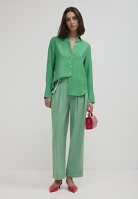 Woman wearing a green buttoned shirt and light green trousers holding a small red handbag, standing against a plain light background.