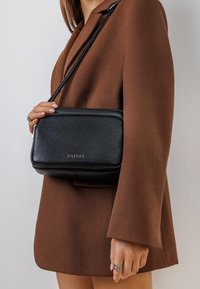 Czarna skórzana torba na ramię o teksturowanym wykończeniu, prostokątnym kształcie i złotym logo. Nosi się ją z brązowym żakietem.