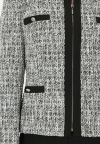 Teksturowana czarno-biała kurtka z srebrnymi elementami, zamek błyskawiczny z przodu, dwie kieszenie z klapkami, kontrastujące czarne akcenty na kieszeniach i kołnierzu.