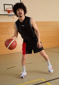 Μαύρη αμάνικη φανέλα με λευκές λεπτομέρειες, μαύρη και κόκκινη βερμούδα, λευκά και πολύχρωμα παπούτσια μπάσκετ. Κρατάει μια μπάλα μπάσκετ μέσα σε ένα γυμναστήριο.