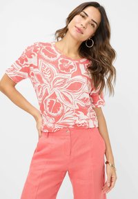 Korallefarbener Kurzarm-Top mit einem auffälligen floralen und blätterartigen weißen Muster; kombiniert mit passenden korallefarbenen Hosen, die eine lockere Passform zeigen.