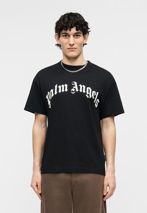 Νέος άνδρας με σγουρά μαλλιά που φοράει μαύρο T-shirt "Palm Angels", αλυσίδα στο λαιμό και καφέ παντελόνι, στέκεται μπροστά σε απλό φωτεινό φόντο.