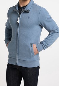 Ljusblå zip-tröja med sidofickor, ribbade ärmslut och fåll, med en liten logotyp på bröstet och en randig inre krage.