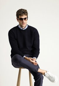 Marine trui, blauw geruite shirt, grijze broek en witte sneakers. Draagt donkere zonnebril, zittend op een houten kruk tegen een lichte achtergrond.