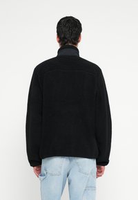 Juoda vilnos striukė su aukštu apykakle ir laisvu siluetu, pasižyminti tekstūros ir siūlių detalėmis, derinama su šviesiai mėlynais džinais.