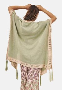 Poncho verde claro de corte holgado, decorado con un ribete de encaje crema y borlas en las esquinas, con una textura de punto suave.