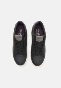 Crne tenisice sa glatkim kožnim gornjim dijelom, teksturiranim akcentima, crnim vezicama i ljubičastim ulošcima označenim s "Wrangler" u sivoj boji. Bijeli gumeni potplat.