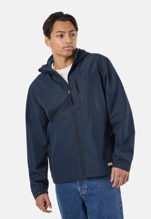 Jonge man die een marineblauw jack met rits en capuchon draagt en een blauwe spijkerbroek, kijkt naar rechts tegen een effen achtergrond.