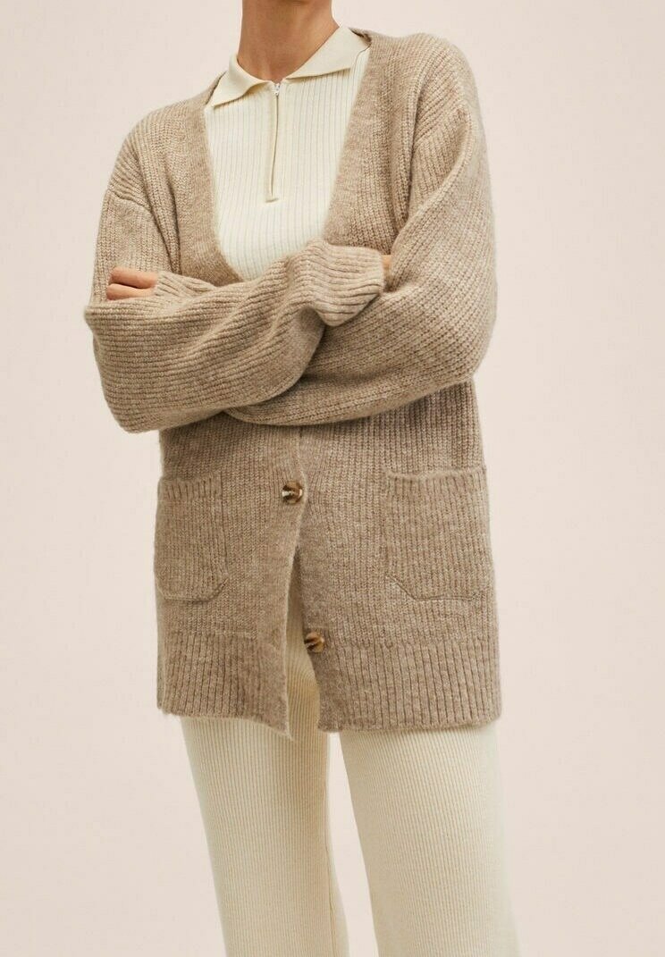 Personne portant un cardigan beige en maille avec des poches devant, par-dessus un haut zippé côtelé crème et un pantalon côtelé assorti, bras croisés.