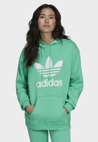 Žalias hoodie, pagamintas iš švelnios medžiagos, su priekinėmis kengūromis kišenėmis, su baltu adidas logotipu ir klasikiniais trimis dryželiais ant rankovių.