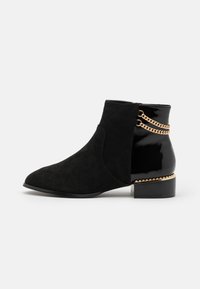 Botines negros con acabado de ante, punta afilada, tacón de charol brillante y detalle de cadena dorada en la parte trasera.